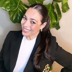 A woman in black jacket and white shirt standing next to green plant.